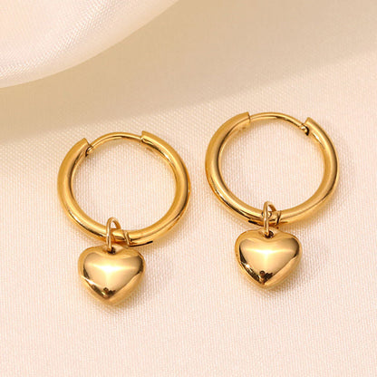 A pair of gold hoop earrings with small polished heart charms, displayed on a soft cream fabric background.