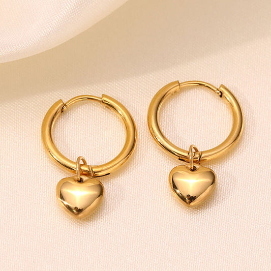 A pair of gold hoop earrings with small polished heart charms, displayed on a soft cream fabric background.